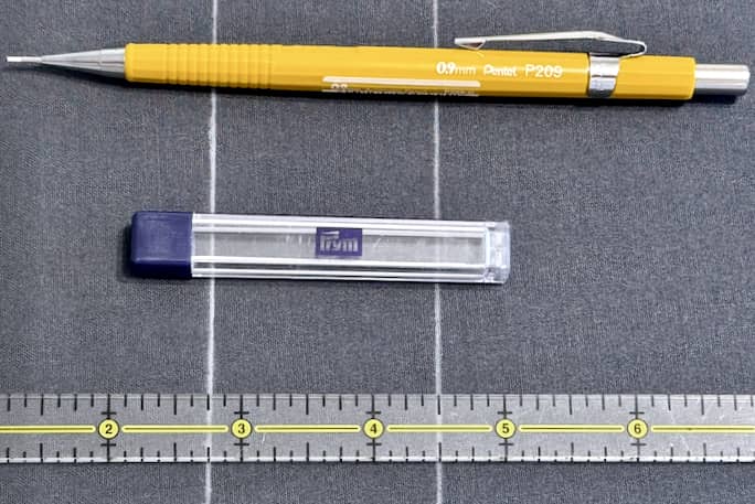 A Pentel brand mechanical pencil loaded with white chalk sits atop grey fabric; a small plastic Prym-branded container containing white chalk refills sits in the centre; a thin clear ruler sits at the bottom of the photo for context.